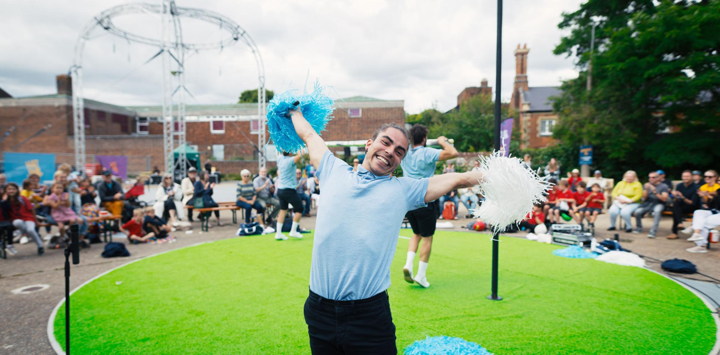 A person dressed in a blue polo top and dark blue shorts waving pom poms and performing outdoors on a green carpet stage