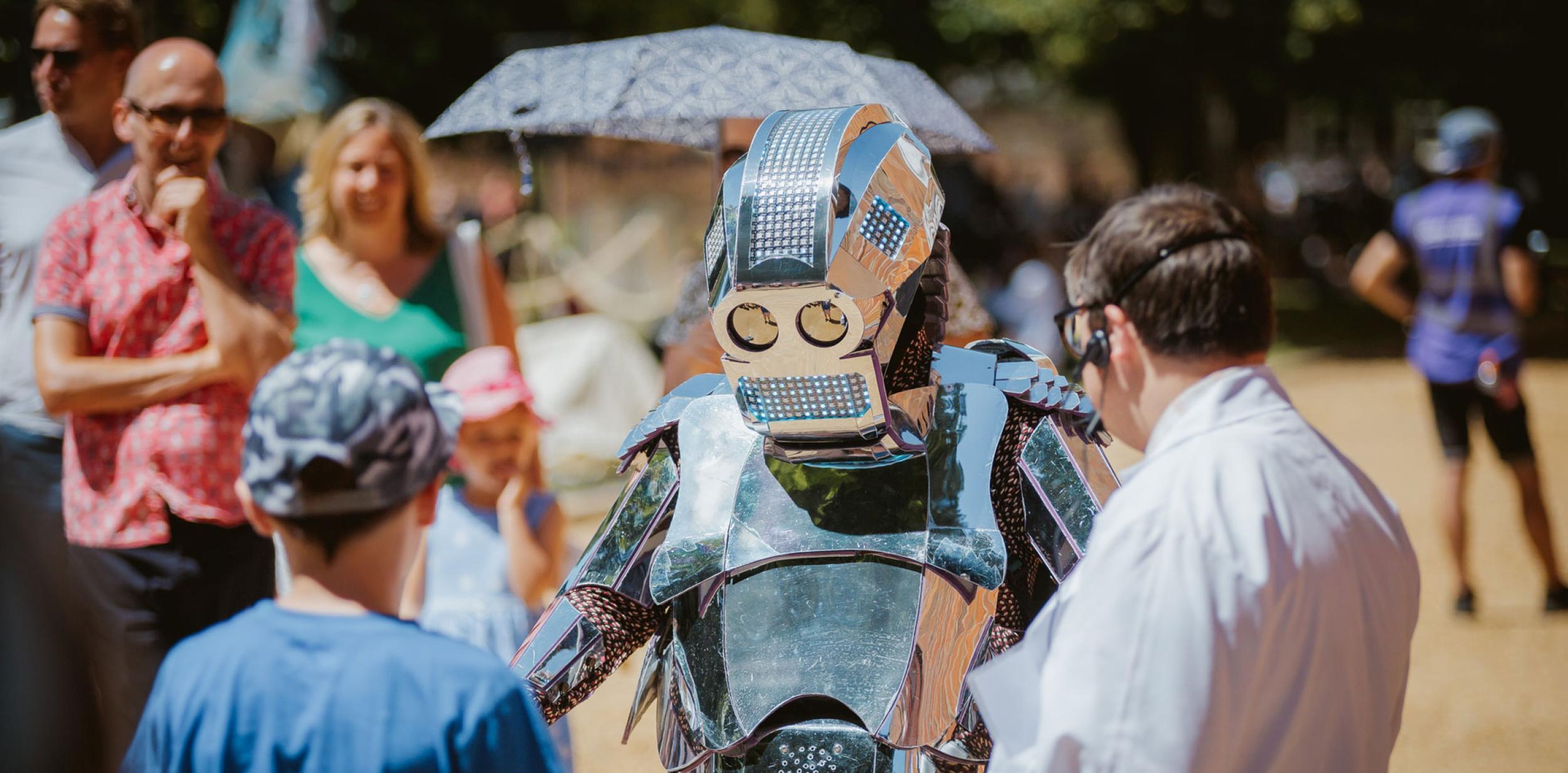 Performer at Hat Fair 2018 Outdoor Arts festival. A life size robot interacting with a child and a person in a lab coat.