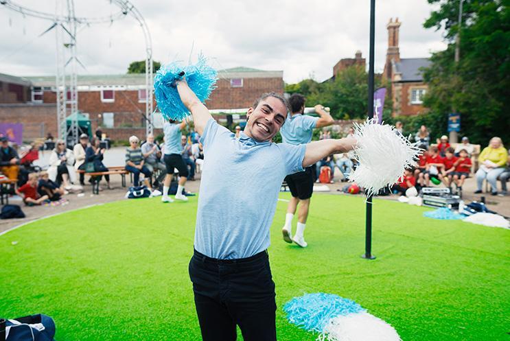 A person dressed in a blue polo top and dark blue shorts waving pom poms and performing outdoors on a green carpet stage